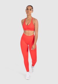 Rode sportbeha met verstelbare banden, gecombineerd met hoge rode leggings, beide gemaakt van soepel, rekbaar materiaal. Gemaakt met witte sneakers.