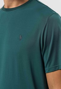 Tmavě zelené tričko s kulatým výstřihem, krátkými rukávy a malým vyšitým logem na hrudi. Hladká, texturovaná tkanina.
