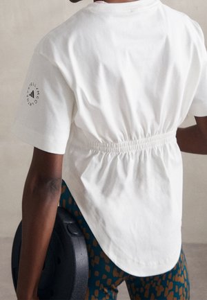 Personne portant un t-shirt blanc cintré dans le dos et des leggings à motifs, tenant une plaque de poids noire dans la main droite.