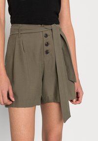 Olivově zelené šortky s vysokým pasem a zavazovacími pásky, s plisovaným designem a třemi tmavými knoflíky vpředu. Lehčí materiál.