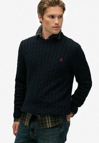 Mann in marineblauem Strickpullover über einem grün-karierten Hemd und blauen Jeans, der zur Seite schaut und eine Hand in der Tasche hat.