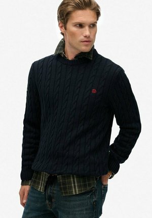 Man in een marineblauwe kabelgebreide trui over een groen geruit overhemd en blauwe jeans, kijkt naar de zijkant met één hand in zijn zak.