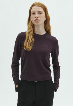 Jeune femme aux longs cheveux roux portant un haut à manches longues violet foncé et un pantalon noir, debout les mains dans les poches devant un fond uni.