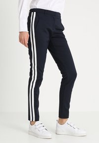 Granatowe spodnie z dwiema pionowymi,białymi paskami; dopasowany krój; wykonane z gładkiego, elastycznego materiału; noszone z białymi sneakersami.