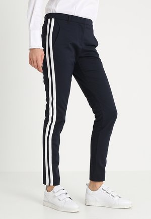 Marineblauwe broek met twee verticale witte streepaccenten; tapering fit; gemaakt van een glad, rekbaar materiaal; gedragen met witte sneakers.