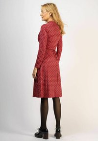 Punainen kietaisumekko geometrisella kuviolla, pitkät hihat ja vyötärölle sidottu nauha. Mallilla on päällään mustat legginsit ja matalakorkoiset kengät.