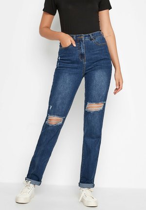 Jean en denim bleu taille haute avec des déchirures usées au niveau des genoux, coupe droite et décoloration subtile. Associé à des baskets blanches.
