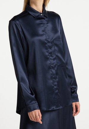Femme portant une chemise en satin bleu marine à manches longues avec une poche poitrine et un col boutonné, associée à une jupe assortie.