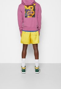 Fialová mikina s grafikou slunečnice a textem, modrá tričko pod ním, žluté šortky, bílé ponožky a zelené tenisky se žlutými akcenty.