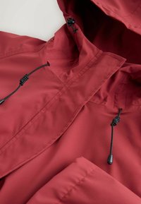 Rdeča vodoodporna jakna z visokim ovratnikom, nastavljivimi črnimi vrvicami in gladko teksturo. Ponaša se z minimalističnim dizajnom in šivi.