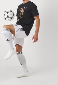 Musta t-paita, jossa on leijonagrafiikka ja vaaleanpunaiset aurinkolasit. Yhdistetty valkoisiin shortsihousuihin, joissa on mustat raidat, ja valkoisiin jalkapallokengiin. Pitelee jalkapalloa.