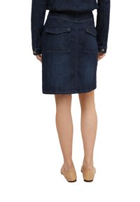 Falda de mezclilla oscura con un diseño ajustado, bolsillos traseros y costuras visibles. Se muestra desde atrás, combinada con zapatos beige sin cordones.