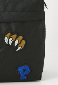 Črn nahrbtnik iz blaga z rjavimi kremplji in modrimi črkami "P" na njemu. Teksturirana površina z vezenimi vzorci in zadrgo.