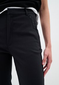 Black tailored trousers with a smooth texture, featuring side pockets and a slim fit design. A zipper accent is visible on one side.