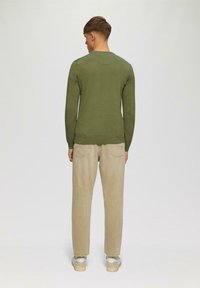 Olivgrüner, langärmliger Strickpullover mit Rundhalsausschnitt, kombiniert mit hellbeigen, schmal zulaufenden Hosen und weißen Schuhen.