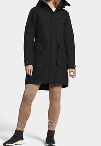 Femme portant un long manteau noir à capuche avec des poches avant, des leggings beige côtelés et des baskets noires à enfiler avec des semelles blanches.