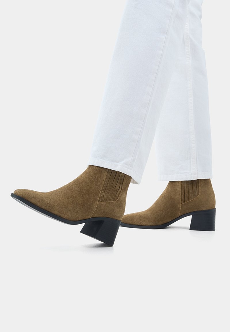 Bottines en daim marron à talons blocs bas portées avec un pantalon blanc, vues de côté sur un fond clair uni.