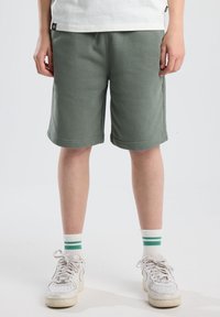 Pantaloni corti in cotone verde con vestibilità relaxed, dotati di un elastico in vita e tasche laterali. Indossati con sneaker bianche e calzini a righe.
