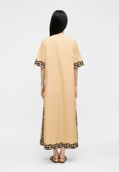 Femme aux longs cheveux noirs portant une robe beige longue jusqu'aux chevilles avec des motifs circulaires noirs sur les manches, l'ourlet et les côtés, vue de dos, debout.