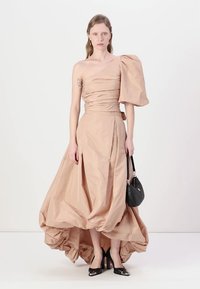 Beige, one-shoulder dress with a gathered bodice, flowing fabric, and a high-low hem. Paired with black pointed heels and a small handbag.