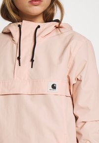 Jasnoróżowy polar z kapturem z czarnymi sznurkami, przednią kieszenią i białą łatą z logo Carhartt. Gładka tekstura, rękawy do kostek.