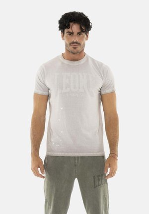 Man met donker haar met een lichtgrijs "LEONE 1947" t-shirt en olijfgroene short staand tegen een witte achtergrond.