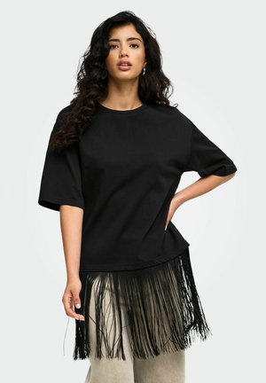 Femme aux cheveux bouclés foncés portant une chemise noire ample avec de longues franges à l'ourlet et un pantalon clair, debout, une main sur la hanche.
