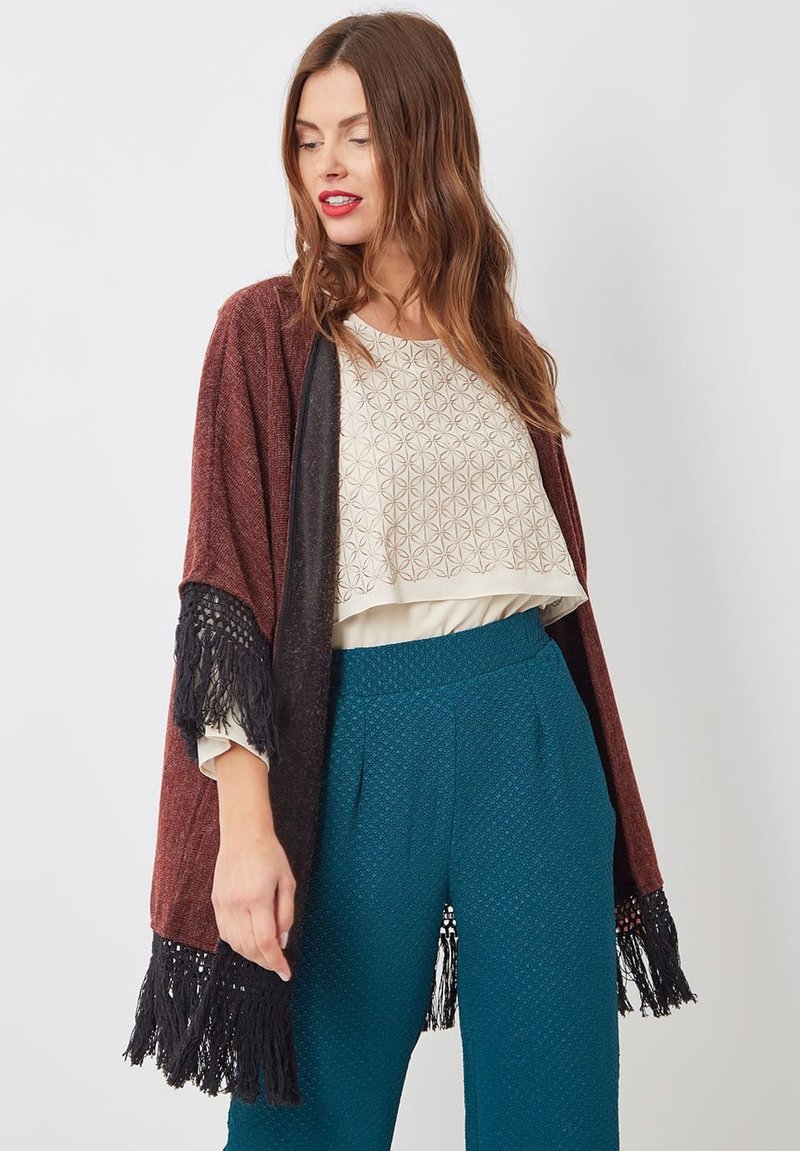 Mujer vistiendo una blusa crema con estampado, pantalones en tono teal texturizados y un chal marrón burdeos con flecos negros, mirando ligeramente hacia su izquierda.