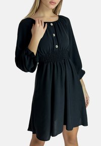 Schwarzes Kleid aus leichtem Stoff, mit geschmocktem Taillengürtel, langen Ärmeln und Knopfdetail am Ausschnitt. Sauberes, einfaches Design.