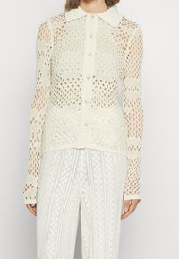 Strickjacke in Creme mit Kragen, Knopfverschluss und Lochmuster über dem Körper und den Ärmeln.
