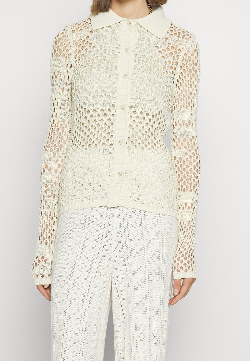 Strickjacke in Creme mit Kragen, Knopfverschluss und Lochmuster über dem Körper und den Ärmeln.