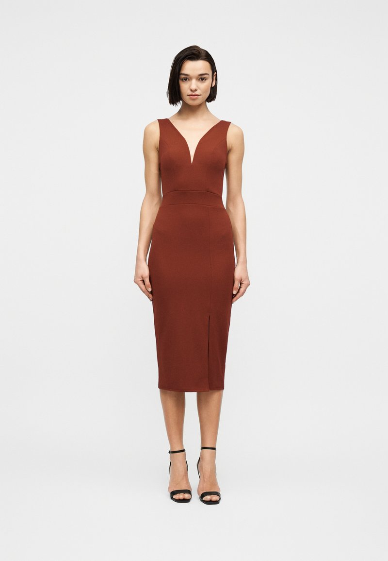 Γυναίκα που στέκεται με αμάνικο φόρεμα μέχρι το γόνατο σε χρώμα σκουριάς, με V-λαιμόκοψη και εμπρόσθια σχισμή, φορώντας μαύρα ψηλοτάκουνα με λουράκι στον αστράγαλο σε λευκό φόντο.