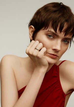 Anillo negro con pequeñas piedras preciosas en una mano, que presenta una piel suave y pálida. El sujeto lleva una blusa roja, drapeada y sin mangas.