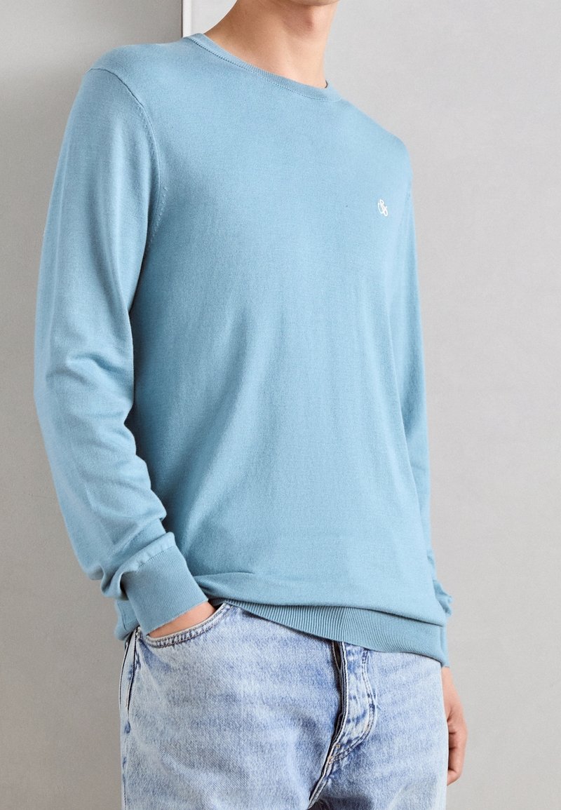 Homme portant un pull à manches longues bleu clair avec un petit logo blanc, associé à un jean denim bleu clair, mains dans les poches.