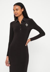 Fekete hosszú ujjú pólóruha rugalmas anyagból, gallérral, gombos előlappal és egy gyémánt alakú logóval a bal mellrészen.