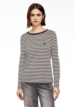 Young woman with straight brown hair wears a black-and-white striped long-sleeve shirt and gray jeans, standing against a white background.