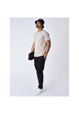 Man draagt een zwarte bucket hat, beige grafisch T-shirt, zwarte cargobroek, witte sneakers, houdt een zwarte clutch vast, staat op een effen achtergrond.