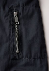 Marinblått tyg med en glänsande silvermetalldragkedja vid ficköppningen, har en slät yta och platta sömmar.