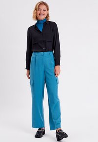 Chemise noire à bouton, pantalon cargo bleu taille haute avec poches latérales, mocassins noirs épais, se tenant devant un fond neutre.
