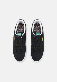 Black suede sneakers with white rubber soles, black laces, and green logo accents. Interior features a patterned insole.