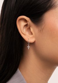 Silberne Creolen mit einem hängenden Kreuzdesign, besetzt mit kleinen klaren Edelsteinen, die am Ohr eines Modells vor glatter Haut präsentiert werden.