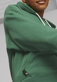Zielona bluza z kapturem, wykonana z miękkiego materiału, z kapturem na sznurku, ściągaczami na rękawach i małą czarną metką z logo po boku.