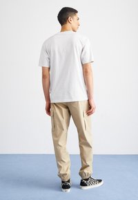 Hellgraues Baumwoll-T-Shirt, beige Cargo-Hose mit Seitentaschen, elastischen Bündchen, schwarz-weiß karierte Sneaker. Stehende Pose, Rückansicht.