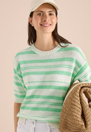 Jeune femme souriante portant un pull rayé vert et blanc et une casquette blanche, tenant un tapis ou une couverture tissée roulée.