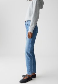 Persona che indossa jeans aderenti blu chiari, felpa grigia e ballerine nere con cinturino, in piedi di lato su uno sfondo neutro.
