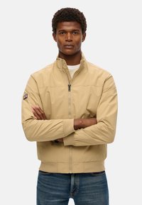 Beige letvægtsjakke med høj krave, lynlås foran og ribstrikkede manchetter. Har et logo-patch på venstre ærme. Båret over en hvid skjorte.