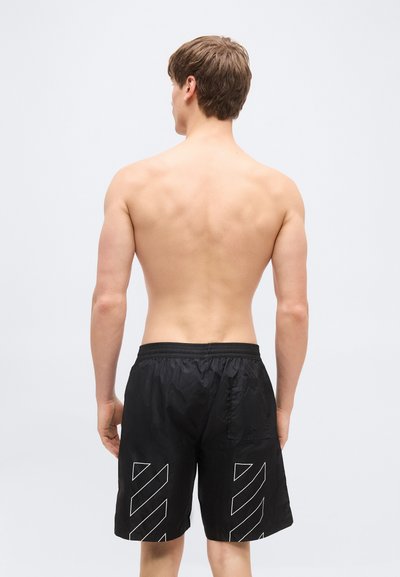 Jeune homme debout, torse nu, de dos, portant un short noir avec des motifs de bandes diagonales blanches à l'arrière de chaque jambe.
