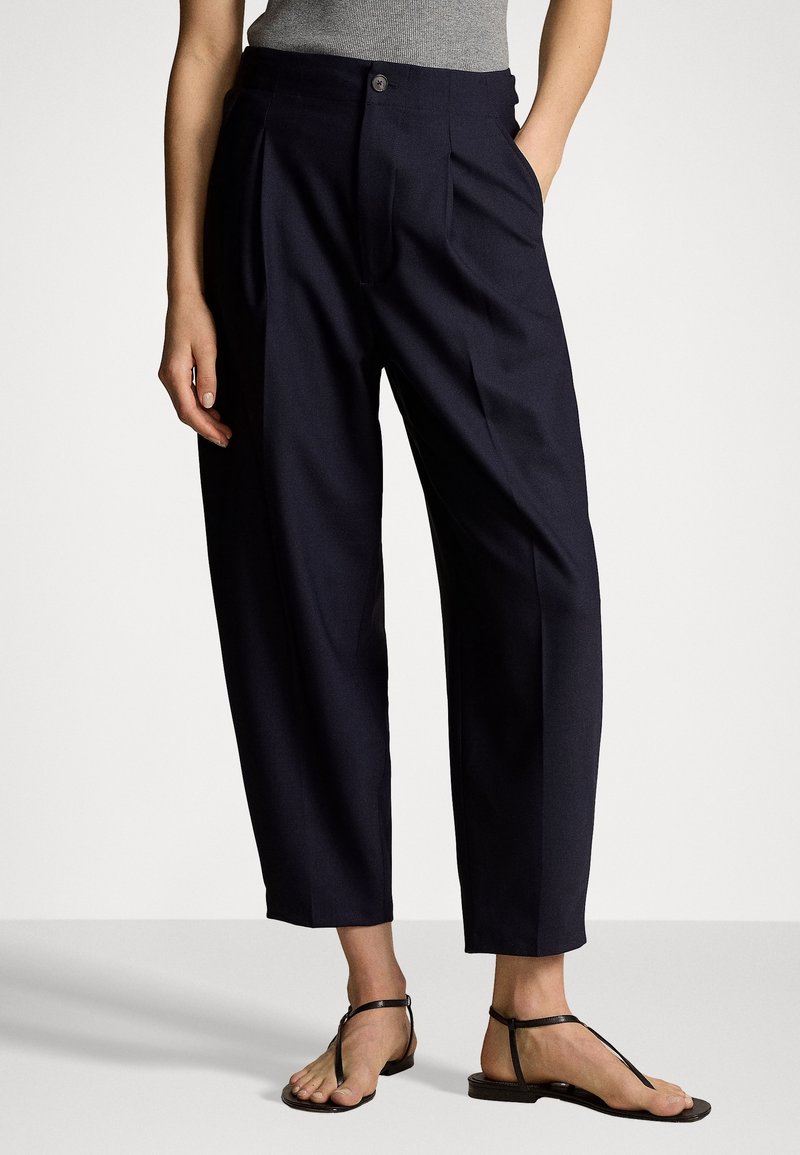Femme portant un pantalon noir sur mesure, court, avec des plis, un top gris sans manches et des sandales plates noires avec des lanières à la cheville.