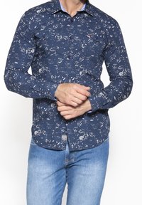 Námořnická modrá košile na knoflíky s bílými skvrnami, dlouhými rukávy, límečkem a dvěma kapsami na hrudi. Obsahuje světle modré detaily.