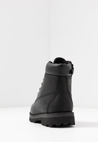 Bota de cuero negro con un collar acolchado, diseño con cordones y suela de goma texturizada. Presenta un logo en el costado y una forma minimalista y aerodinámica.
